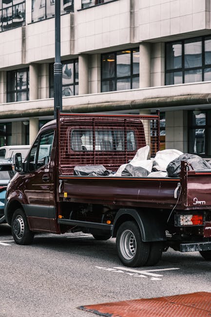 A small dark red flatbed truck parked on a city street in front of a modern multi-storey building with large rectangular windows and concrete facades. The truck's cargo area is loaded with various types of rubbish, including black plastic refuse bags, white plastic bags, and other miscellaneous waste items. The waste appears to be a mixture of household rubbish and debris, casually piled in the open bed of the truck. The vehicle is positioned near a car park area, with a white van partially visible behind it. A black streetlamp is situated in the foreground, slightly to the left, casting a subtle shadow. The surrounding environment suggests an urban setting where private waste collection or on-site clearance services, such as those provided by Waste Disposal Bow, might operate as an alternative to council rubbish collection. The scene is captured during daylight, with natural light highlighting the textures of the waste materials and the vehicle's metallic finish, emphasizing the practical nature of rubbish removal services in busy city environments.