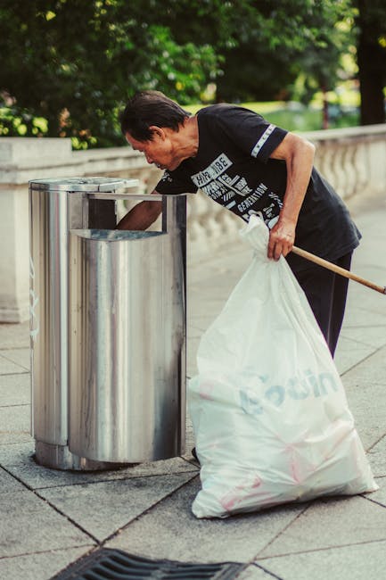 A middle-aged man with dark hair, dressed in a black t-shirt with white text and dark trousers, is seen disposing of rubbish into a metallic recycling bin on a paved outdoor area. He is bent over, using his right hand to place waste into the open top of the cylindrical, stainless steel bin, which has a smooth, reflective surface. In his left hand, he holds a large white plastic bag filled with waste, with the bag's sides wrinkled and gathered at the top. The background features a low stone wall with a concrete cap and lush green trees, creating a shaded environment with dappled sunlight. This scene illustrates an individual participating in private rubbish disposal or on-site waste clearance, emphasizing the importance of proper waste handling outside of municipal collection. The setting's clean, organized appearance aligns with professional rubbish removal services such as those offered by Waste Disposal Bow, supporting alternative waste disposal methods like independent collection or commercial rubbish clearance.