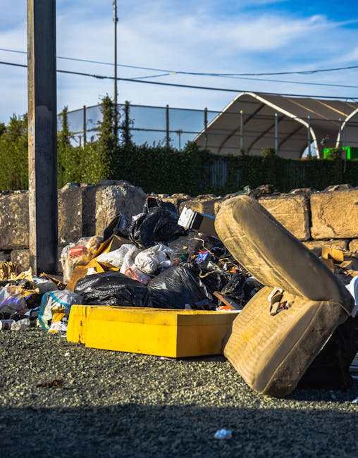 A small dark red flatbed truck parked on a city street in front of a modern multi-storey building with large rectangular windows and concrete facades. The truck's cargo area is loaded with various types of rubbish, including black plastic refuse bags, white plastic bags, and other miscellaneous waste items. The waste appears to be a mixture of household rubbish and debris, casually piled in the open bed of the truck. The vehicle is positioned near a car park area, with a white van partially visible behind it. A black streetlamp is situated in the foreground, slightly to the left, casting a subtle shadow. The surrounding environment suggests an urban setting where private waste collection or on-site clearance services, such as those provided by Waste Disposal Bow, might operate as an alternative to council rubbish collection. The scene is captured during daylight, with natural light highlighting the textures of the waste materials and the vehicle's metallic finish, emphasizing the practical nature of rubbish removal services in busy city environments.