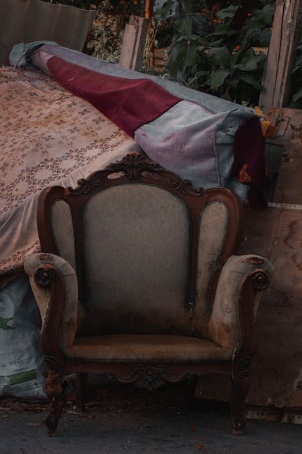 In an indoor or outdoor storage area, a vintage upholstered armchair with a wooden carved frame and light-colored fabric seat and backrest is positioned directly in front of a large pile of various items, including a rolled-up fabric or blanket with grey and burgundy panels. The surrounding environment features wooden and possibly greenhouse or garden structures with visible greenery and foliage in the background, suggesting a setting where household or garden waste may be temporarily accumulated before removal. The chair appears old, with some signs of wear, and is situated next to an assortment of fabric, cardboard, and other miscellaneous objects, indicating a collection of furniture and soft furnishings awaiting disposal or collection by a waste removal service such as Waste Disposal Bow. The scene's natural lighting hints at daytime, and the overall composition aligns with private or non-standard waste handling activities typical in alternative rubbish removal contexts.