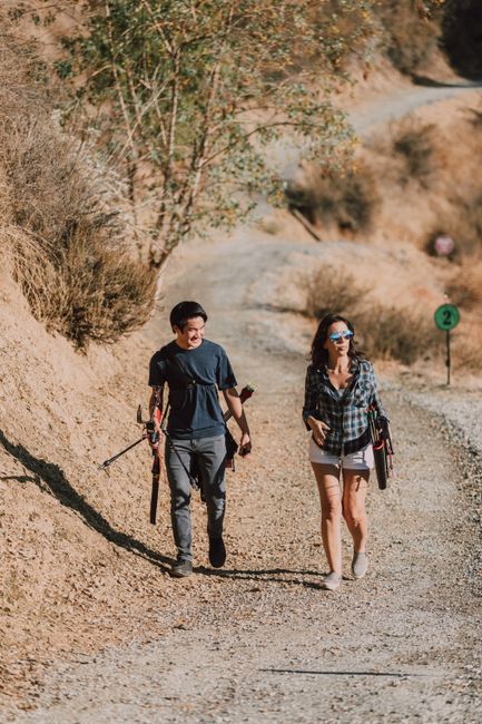 A young man and woman walk along a dusty, uneven trail in a dry, arid landscape with sparse vegetation, including small shrubs and a few trees. The man, on the left, has short dark hair and wears a dark t-shirt, gray trousers, and gloves, carrying a long-handled tool in one hand and a small black bag slung over his shoulder, suggesting involvement in outdoor work or an activity like hiking. The woman, on the right, has long dark hair, wears sunglasses, a checkered shirt, and light-colored shorts, holding a laptop case in one hand. The background features rocky, beige-colored terrain with rolling hills and scattered bushes, under a clear bright sky. The scene is lit by natural sunlight, casting soft shadows on the ground. The setting appears remote and natural, with no visible buildings or infrastructure, and the pathway they walk on is narrow and winding, indicating an outdoor recreational or conservation area where private or alternative waste management practices, such as on-site clearance, might be relevant, indirectly connected to waste disposal activities managed professionally. This image is related to outdoor activities, possibly involving terrain maintenance or environmental management. The focus is on the individuals engaging in their activity amidst the natural environment, with no visible waste or rubbish present.