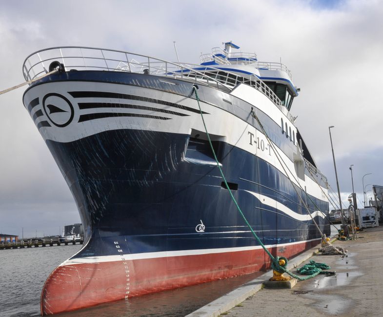 A large motor yacht docked at a port, with a navy blue hull featuring white and black stripe accents along the sides. The bow is prominently visible, showing a streamlined shape, with a white railing running along the top deck. The yacht's lower section is painted in a rusty red color, indicating the waterline, with water streaks visible on the hull's surface. The vessel is secured to the concrete dock by thick green ropes tied to sturdy mooring posts. The surrounding environment includes a cloudy sky overhead, with additional boats faintly visible in the background, along with parked vehicles and port equipment to the side, indicating a busy dockside area. The scene appears to be during daytime with natural lighting highlighting the shiny finish of the yacht's exterior, which is characteristic of private vessels used for leisure or special occasions.