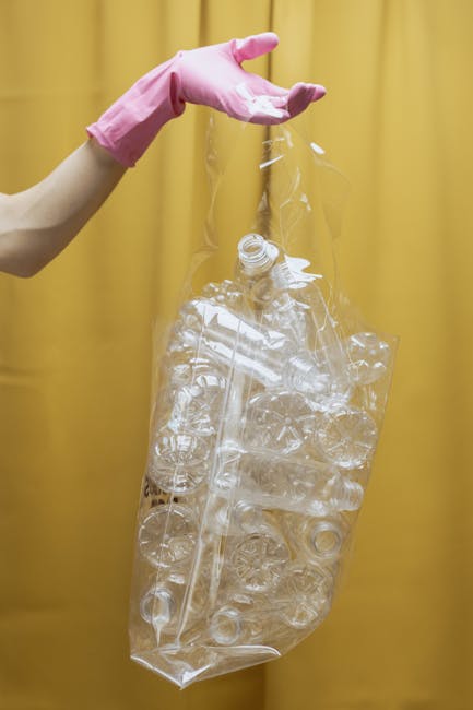 A close-up photograph features a person's hand wearing a light pink rubber glove holding a large, transparent plastic bag filled with numerous empty, clear plastic bottles of various sizes and shapes. The bottles are tightly packed and appear to be used for beverages, with some showing faint residual liquid or labels. The bag is open at the top, hanging downward, with the bottles visibly protruding from the opening. The background consists of a plain, solid yellow curtain or wall, providing a neutral contrast to the clear plastic and the pink glove. The image highlights the process of collecting recyclable plastic waste, indicating an environmental or waste management context, possibly related to rubbish removal or recycling services offered by Waste Disposal Bow. The scene emphasizes the importance of proper plastic disposal and sorting in waste management activities, aligning with independent collection or on-site clearance services.