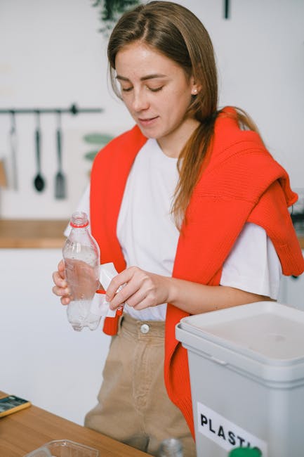 A young woman with long brown hair, dressed in a white T-shirt and an orange sweater draped over her shoulders, is standing indoors in a kitchen or utility area. She is holding a clear plastic water bottle in her left hand, with her right hand opening or sealing the cap. In the background, there is a wall-mounted rack with black kitchen utensils hanging, and a wooden countertop is visible. On the right side of the image, there is a grey plastic container labeled 'PLASTIC,' indicating its use for sorting waste for recycling or disposal. The scene has natural lighting and an organized environment, typical of a household setting engaged in waste sorting or rubbish management, reflecting the practical steps involved in private rubbish clearance or recycling, as supported by waste disposal services such as Waste Disposal Bow.